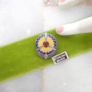 May include: A vintage sterling silver pin and a small rectangular tag with the text "STERLING SILVER". The pin has a circular design with a yellow center, blue and white border, and silver wings. The items rest on a green velvet ribbon.