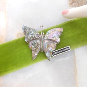 May include: A sterling silver butterfly brooch with iridescent abalone wings rests on a green velvet ribbon. The brooch has detailed antennae and a small tag that reads "STERLING SILVER". The background is a white and beige marble surface.