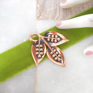 May include: A copper-toned metal brooch shaped like a cluster of three leaves, each featuring a central vein with small raised spherical accents. The metal has a polished finish with darkened, oxidized recessed areas to provide contrast and depth.