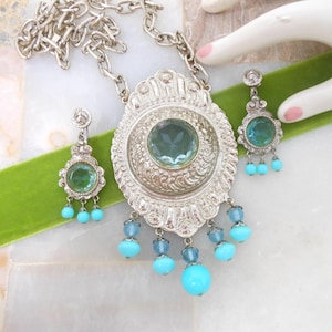 May include: A silver necklace with a large, ornate pendant featuring a light blue gemstone. The pendant is surrounded by smaller turquoise beads. Matching earrings with light blue gemstones and turquoise beads are also included.