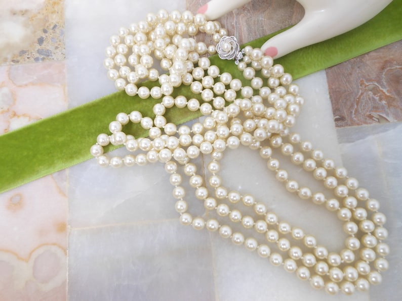 May include: A long strand of white faux pearl necklace with a silver clasp. The necklace is draped over a green velvet ribbon.