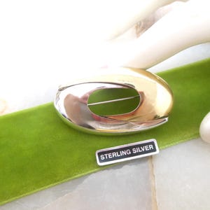 May include: A silver and gold oval-shaped brooch with a cutout in the center. The brooch is labeled "Sterling Silver".