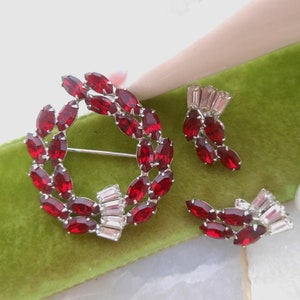 May include: A red and clear rhinestone jewelry set featuring a wreath-shaped brooch and a pair of earrings. The brooch is made of a silver-toned metal and features a circle of red marquise-shaped rhinestones. The earrings are also made of silver-toned metal and feature a cluster of red marquise-shaped rhinestones.