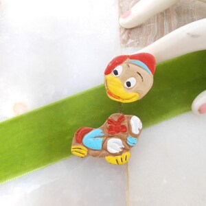 May include: A ceramic pin of a cartoon duck with a red, blue, and white hat. The duck is wearing a blue and white shirt and has a red bow tie. The pin is on a green surface.