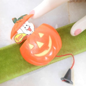 May include: Orange Halloween-themed toy with a smiling jack-o'-lantern face and a ghost popping out. The ghost has the word "Boo!" on it. A black cone is attached by a red string. The toy is on a green ribbon.