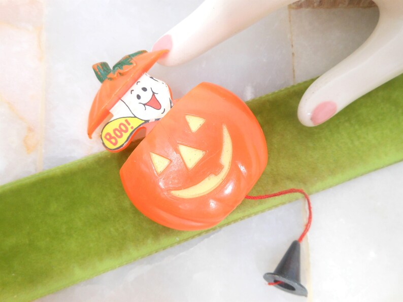 May include: Orange Halloween-themed toy with a smiling jack-o'-lantern face and a ghost popping out. The ghost has the word "Boo!" on it. A black cone is attached by a red string. The toy is on a green ribbon.