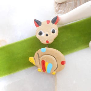 May include: A whimsical, handmade cat-shaped pin with a snail-like body. The cat's head is tan with black ears and red accents. The snail body is tan with blue, red, and yellow details. The pin is set against a green velvet ribbon.