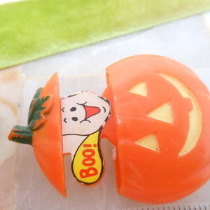 May include: Orange plastic Halloween jack-o'-lantern with a smiling ghost popping out. The ghost has a red mouth and a yellow speech bubble that says "Boo!". The jack-o'-lantern has a green stem.