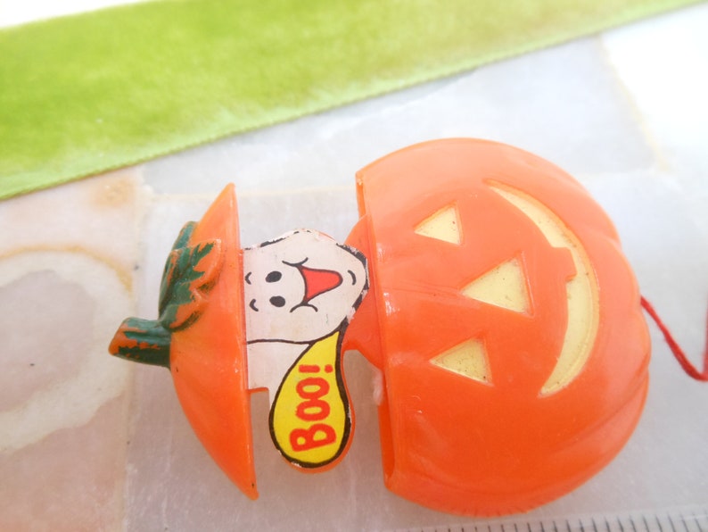 May include: Orange plastic Halloween jack-o'-lantern with a smiling ghost popping out. The ghost has a red mouth and a yellow speech bubble that says "Boo!". The jack-o'-lantern has a green stem.