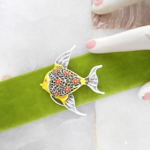 May include: A yellow and silver fish-shaped brooch with orange accents. The brooch is set on a green velvet ribbon. The fish has a detailed design with a lattice pattern and is set against a white background.