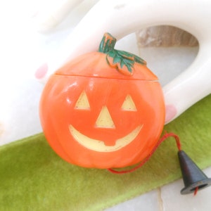 May include: Orange plastic jack-o'-lantern candy container with a smiling face and a green stem. The lid is removable. A black witch hat is attached with a red string. A festive Halloween decoration.