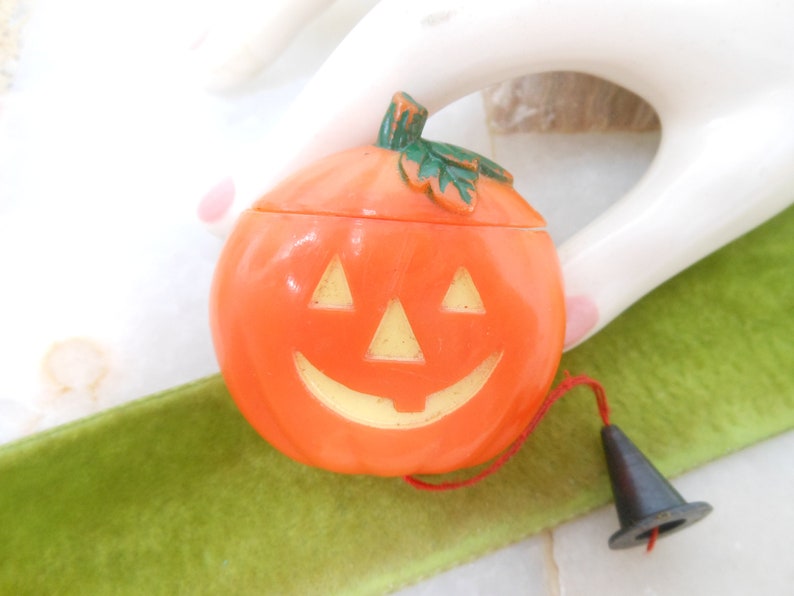 May include: Orange plastic jack-o'-lantern candy container with a smiling face and a green stem. The lid is removable. A black witch hat is attached with a red string. A festive Halloween decoration.