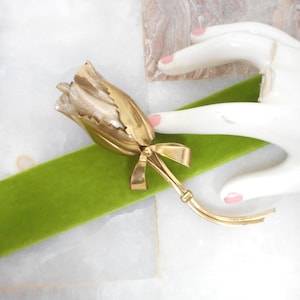 May include: A gold-toned tulip brooch with a bow detail. The brooch is set against a green velvet ribbon and a white marble surface. The tulip's petals have a textured appearance.