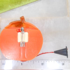 May include: A vintage orange plastic pumpkin-shaped pin with a stem and a black cone-shaped object attached by a red string. The pin has a safety clasp on the back.