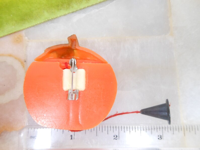 May include: A vintage orange plastic pumpkin-shaped pin with a stem and a black cone-shaped object attached by a red string. The pin has a safety clasp on the back.