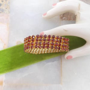 May include: A gold-toned bracelet with rows of small, red gemstones. The bracelet is flexible and appears to be made of multiple connected segments. The gemstones are closely set, creating a continuous band of color. The bracelet is displayed on a white hand.