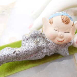 May include: A silver and blue ceramic figurine of a sleeping elf with closed eyes. The elf is lying on a green velvet surface.