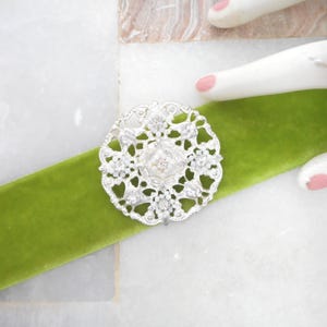 May include: A silver-toned filigree brooch with a central clear stone is displayed on a vibrant green velvet ribbon. The brooch features an intricate floral design. The background is a white and gray marble surface.