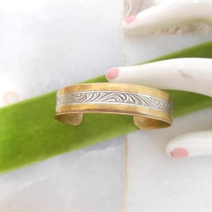 May include: A gold-toned cuff bracelet with a silver-colored, engraved central band featuring a swirling pattern. The bracelet is displayed on a green velvet ribbon and a white surface.