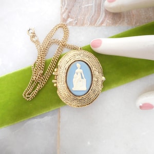 May include: Gold-tone oval locket necklace with a light blue cameo of a woman. The locket has a textured border and hangs on a delicate chain. The necklace rests on a green velvet ribbon.