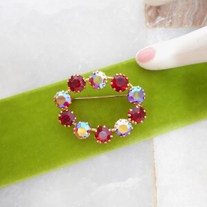 May include: Oval-shaped gold-tone brooch with a ring of red and iridescent crystals. The brooch is set on a bright green velvet ribbon. A hand with a painted fingernail is in the background.