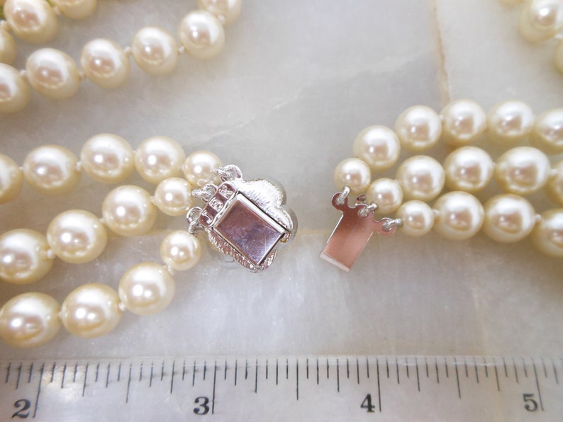 May include: A three-strand pearl necklace with a silver clasp. The necklace is measured against a ruler, showing it is approximately 18 inches long.