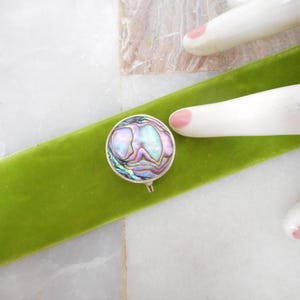 May include: A circular brooch featuring a polished abalone shell inlay with iridescent swirls of purple, blue, and green. The shell is set within a simple silver-toned metal frame, showcasing a smooth, rounded surface and a small metal pin mechanism.