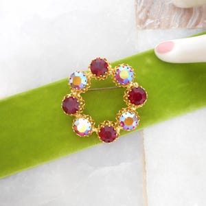 May include: A gold-toned brooch in a circular wreath design, adorned with alternating red and iridescent crystals. The brooch is set against a vibrant green velvet ribbon.