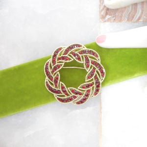 May include: A gold-toned brooch with a woven design, encrusted with red rhinestones. The brooch is circular and rests on a bright green velvet ribbon. The background is a marble surface.