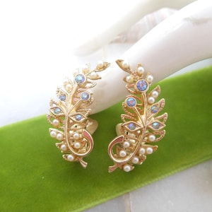 May include: A pair of gold tone earrings shaped like fern leaves with iridescent blue rhinestones and white pearl accents.