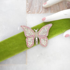 May include: A rhinestone butterfly brooch with pink and clear crystals, set on a green velvet ribbon. The butterfly has a silver-tone metal frame. The brooch is a decorative accessory.