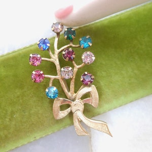 May include: A gold tone brooch with a bouquet of colorful rhinestones. The brooch features a bow at the bottom and a cluster of rhinestones in shades of pink, blue, green, and clear.