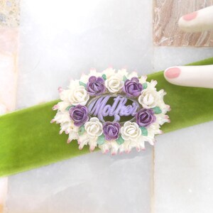 May include: A floral brooch with the word "Mother" in the center, surrounded by white and purple roses. The brooch is set on a bright green velvet ribbon, against a white and marble background.