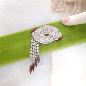 May include: A vintage brooch featuring a dazzling arrangement of clear rhinestones. The brooch has a unique shape with a cascade of dangling rhinestones and accents of red stones. It rests on a vibrant green velvet surface.
