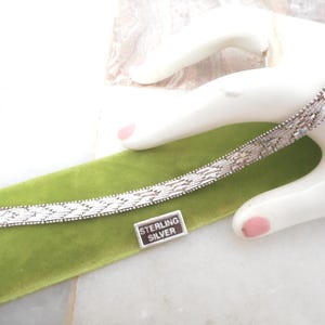 May include: A silver-toned bracelet featuring a woven, textured link pattern. The bracelet includes a metal tag with the text STERLING SILVER engraved in white lettering. It has a secure clasp mechanism at one end and a polished, metallic finish.