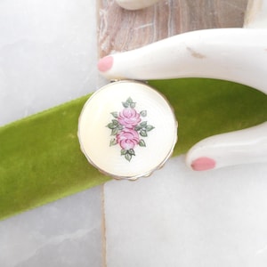 May include: A round, vintage compact with a cream-colored surface and a floral design featuring pink roses and green leaves. The compact has a gold-colored rim and sits on a green velvet ribbon.