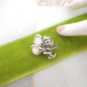 May include: A silver-toned metal brooch shaped like a small winged creature, featuring two smooth, round, off-white cabochon inlays on its wings. The figure has pointed ears and a detailed facial expression, positioned on a bright green velvet ribbon.