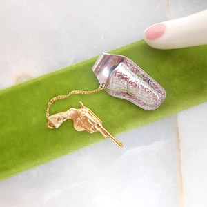 May include: A silver-toned finger ring with an engraved design, connected by a gold chain to a gold-colored revolver charm. The ring is resting on a green velvet surface.