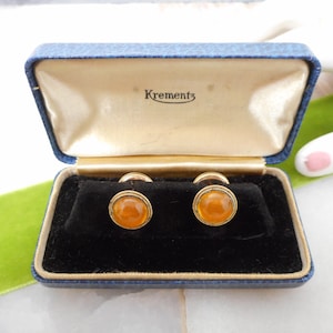 May include: A pair of gold-toned cufflinks with amber-colored centers, presented in a blue box with the brand name "Krementz" printed on the inside lid. The cufflinks are set against a black velvet interior.