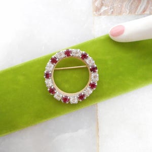 May include: A gold-toned circular brooch with alternating red and clear gemstones. The brooch is set against a bright green velvet ribbon. The pin is a classic jewelry piece.