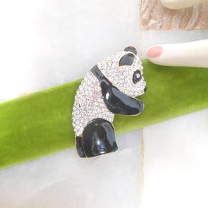 May include: A panda bear brooch with a black and white design. The panda's body is covered in sparkling crystals, with black accents on the ears, arms, and legs. The brooch is set on a green velvet ribbon.