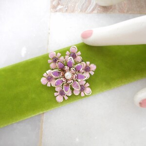 May include: A floral brooch with a pearl center and pink and purple petals is displayed on a green velvet ribbon. The brooch has a gold-colored metal frame. The background is a white marble surface.
