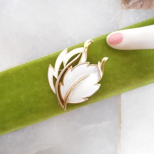 May include: A vintage leaf brooch with white enamel and gold-tone metal accents. The brooch features a stylized leaf design, resting on a green velvet ribbon. The brooch is approximately 2 inches in length.