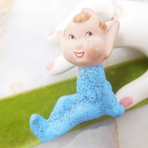 May include: A small ceramic figurine of an elf wearing a blue jumpsuit with a speckled texture. The elf has a pink hat and a smiling face.