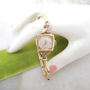 May include: A gold-tone analog wristwatch featuring a square case with a white dial and gold-tone markers. The watch has a delicate link bracelet with decorative scrollwork details at the lugs where the band connects to the watch face.