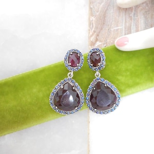 May include: A pair of dangle earrings with a dark red teardrop-shaped stone, surrounded by small blue stones. The earrings have a smaller oval-shaped stone at the top, also surrounded by blue stones. The earrings are set against a green velvet ribbon.
