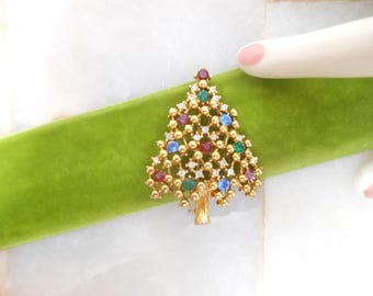 May include: A gold-toned Christmas tree brooch with red, green, blue, and clear gemstone accents. The brooch is set against a bright green velvet ribbon.