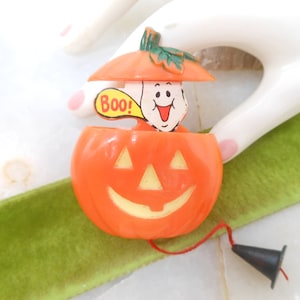May include: Orange plastic Halloween jack-o'-lantern toy with a smiling face and a ghost figure popping out, saying "Boo!". The ghost is wearing a pumpkin hat. A black cone-shaped piece is attached to the toy with a red string.