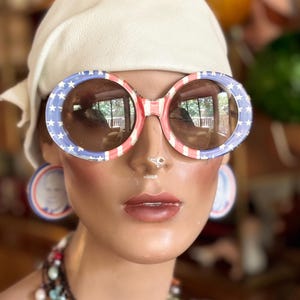 May include: A white headband with a red, white, and blue American flag pattern on a mannequin wearing large round sunglasses with the same pattern. The mannequin is wearing a multi-colored beaded necklace.