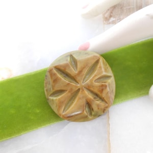 May include: A round, brown button with a carved, starburst design. The button is on a green velvet ribbon.